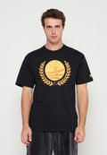 Junger Mann trägt ein schwarzes T-Shirt mit orangefarbenem Lorbeerkranz und rundem Emblem mit einer Figur und Streifen vorne, stehend vor einfachem Hintergrund.