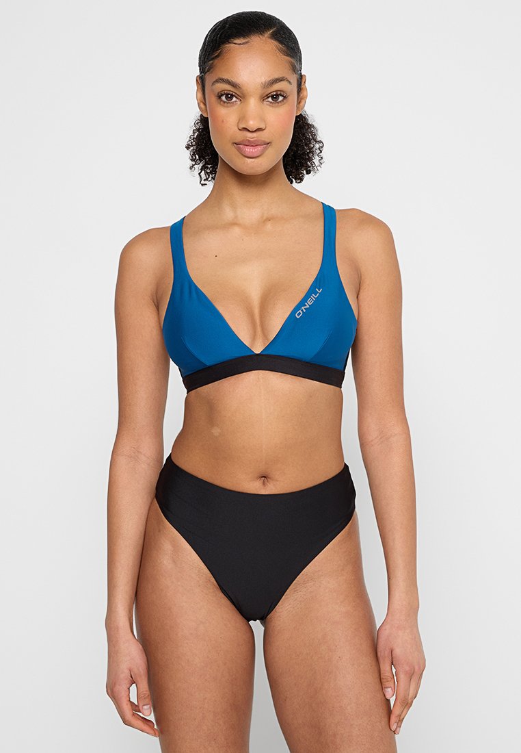 Frau mit lockigem Haar in einem blauen O'Neill-Bikini-Oberteil und einer schwarzen Bikinihose mit hoher Taille, die vor einem schlichten weißen Hintergrund steht.