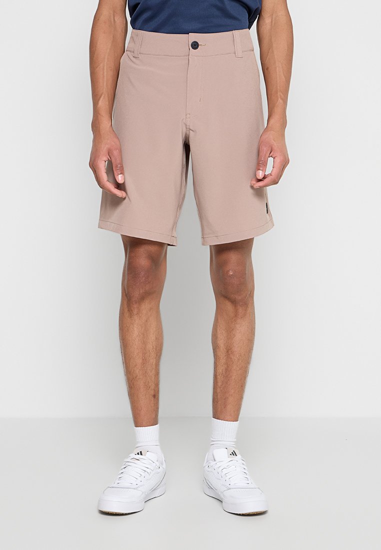 Mann trägt beige knielange Shorts, weiße Socken und weiße Adidas-Sneaker, steht vor einem schlichten hellen Hintergrund.