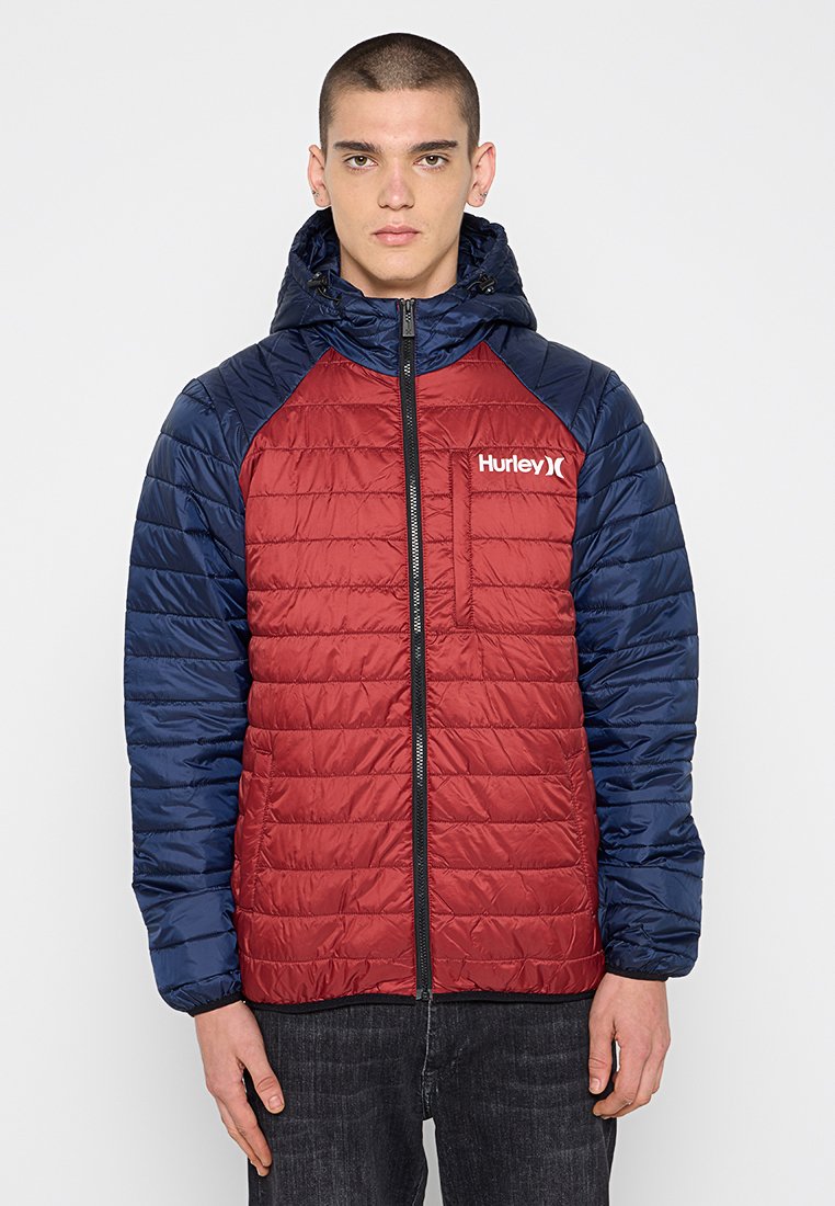 Rote und marineblaue isolierte Jacke mit einem gesteppte Muster, Frontreißverschluss, Kapuze und Logo auf der Brusttasche. Diese Jacke hat lange Ärmel.