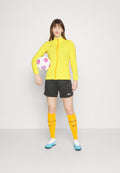 Gelbe sportliche Zip-Jacke, schwarze Shorts, orange Socken mit schwarzen Streifen, hellblaue Fußballschuhe, hält einen mehrfarbigen Fußball.