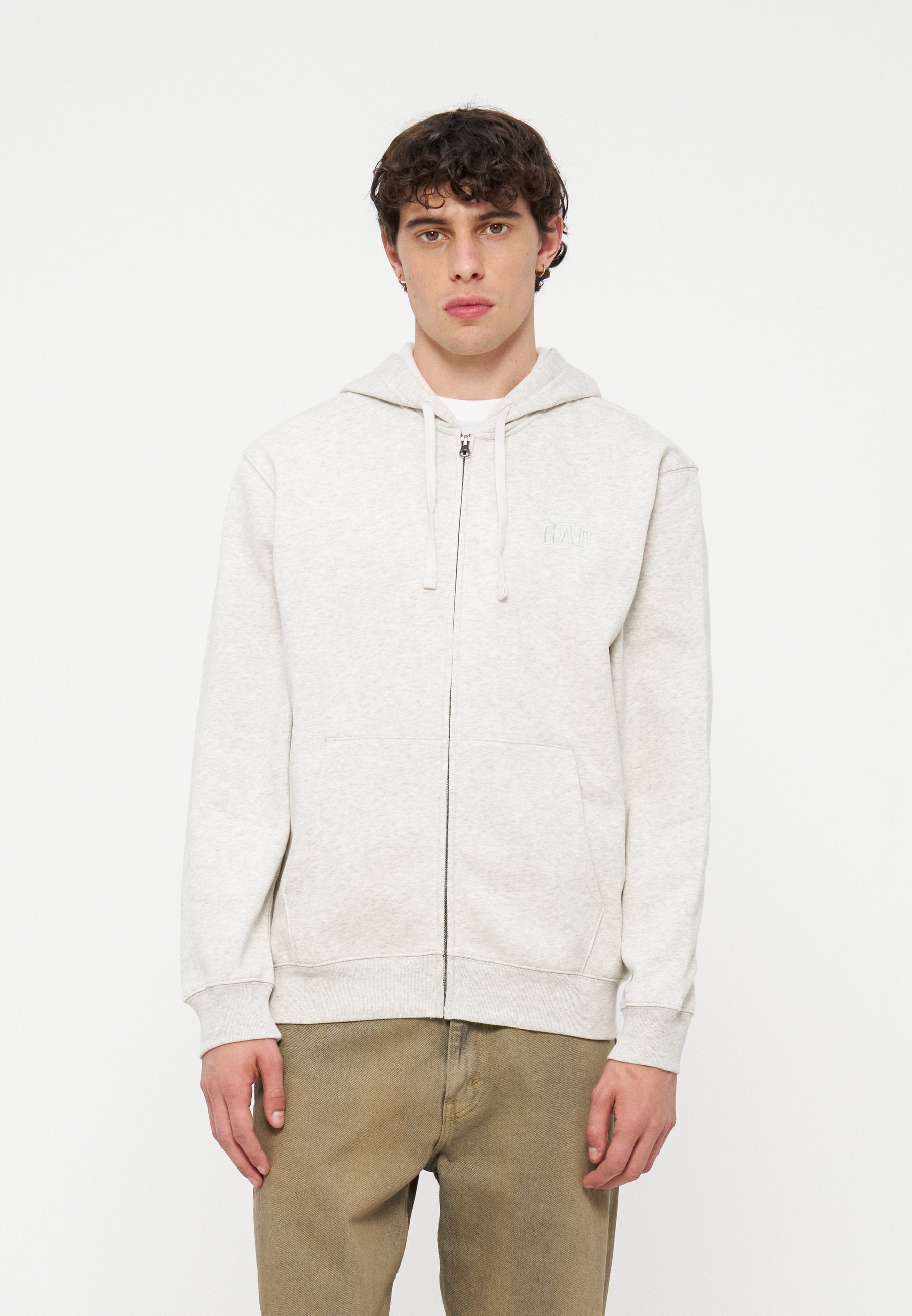 Junger Mann stehend, trägt einen hellgrauen Kapuzenpullover mit Reißverschluss und Fronttaschen sowie beige Hosen vor einem schlichten weißen Hintergrund.
