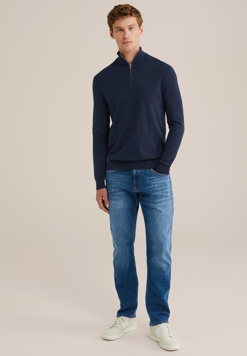 Junger Mann, der mit einer Hand in der Tasche steht, trägt einen navyfarbenen Quarter-Zip-Pullover, blaue Jeans und weiße Sneakers, vor einem beigen Hintergrund.