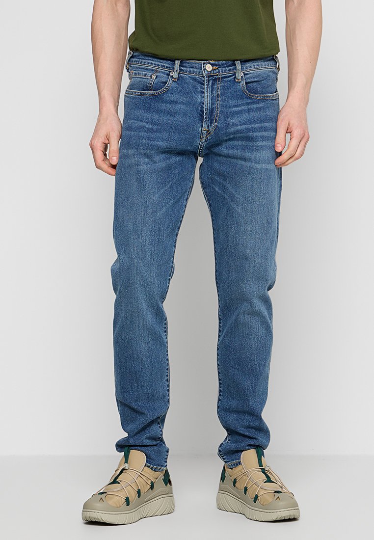 Mann in mittelblauen, geraden Jeans, die mit beigen Sneakers mit grünen Akzenten kombiniert sind, steht vor einem neutralen Hintergrund.