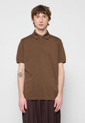 Junger Mann mit kurzen braunen Haaren, der ein braunes kurzärmeliges Poloshirt trägt, vor einem schlichten hellen Hintergrund steht und eine Tätowierung auf seinem linken Unterarm hat.