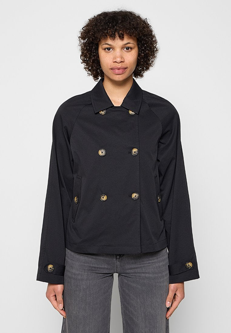 Frau mit lockigem Haar, die eine schwarze, zweireihige Jacke mit Knöpfen und graue Jeans trägt, vor einem hellgrauen Hintergrund steht.