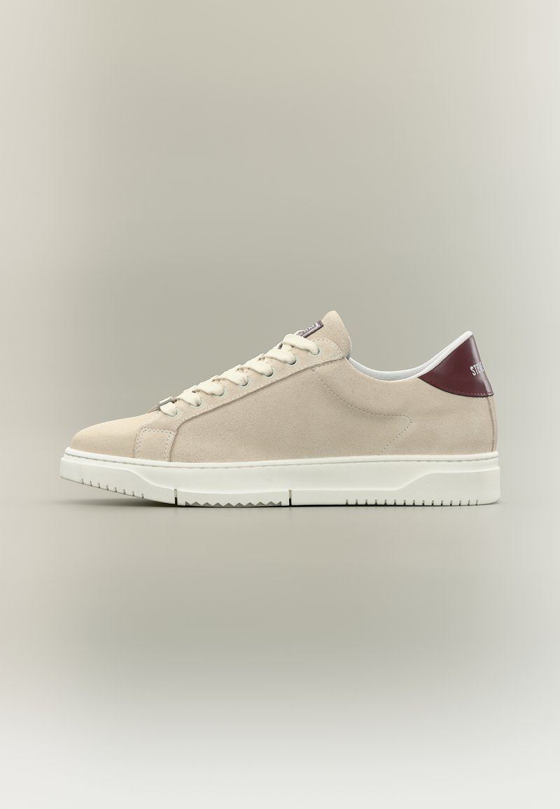 Beiger Wildleder-Sneaker mit niedrigem Schaft, weißer Sohle, beigen Schnürsenkeln und dunkelrotem Fersenlasche mit weißem Textlogo.