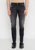 Mann trägt schmal geschnittene ausgewaschene schwarze Jeans und schwarze Schnallenschuhe, steht vor einem einfarbigen hellen Hintergrund.