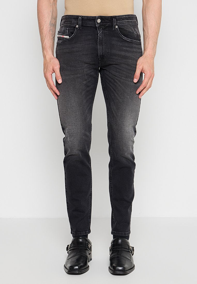 Mann trägt schmal geschnittene ausgewaschene schwarze Jeans und schwarze Schnallenschuhe, steht vor einem einfarbigen hellen Hintergrund.