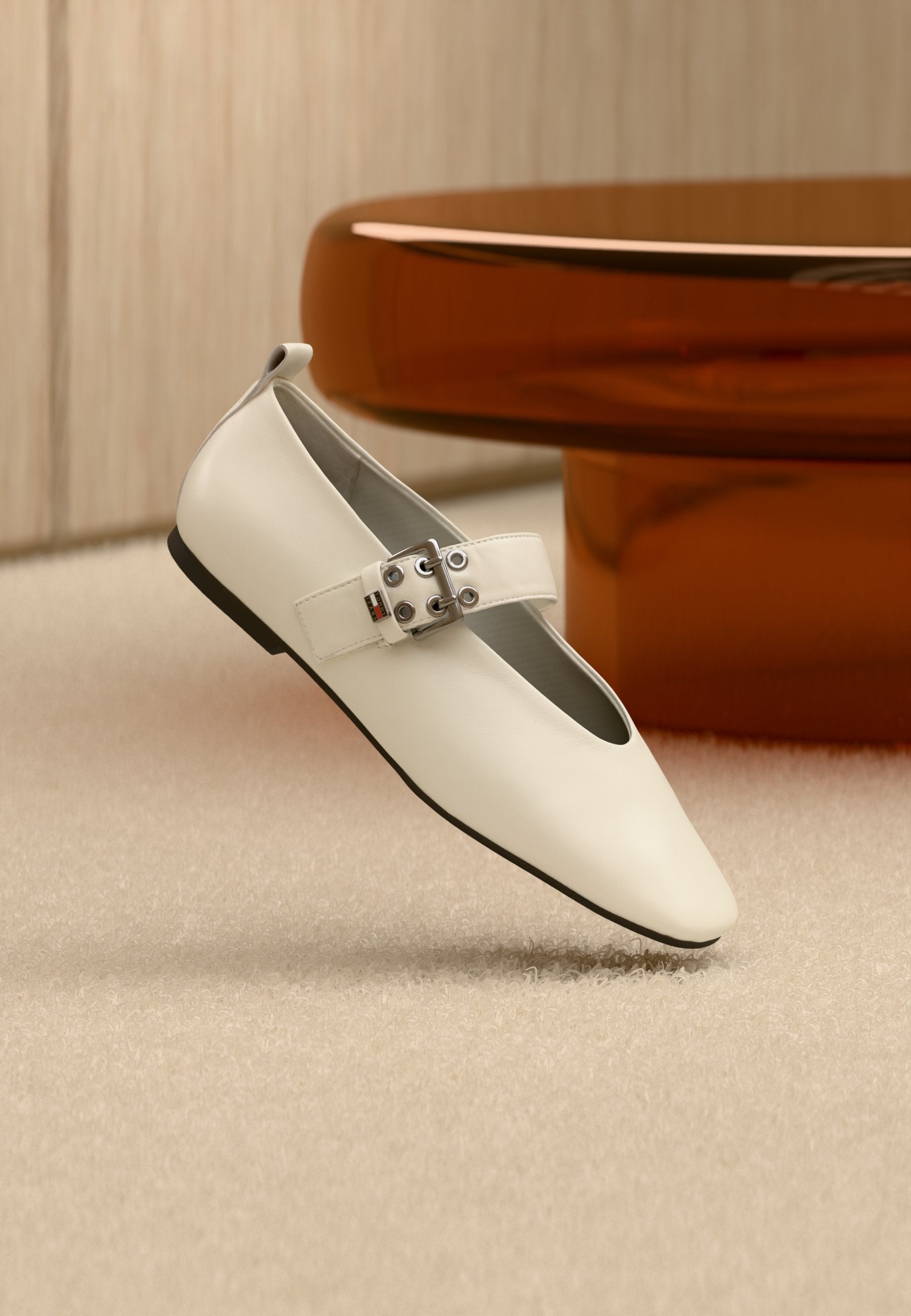 Weißer Mary Jane Ballerina aus Leder mit silberner Schnalle und schwarzer Sohle, auf einem beigen Teppich vor einem braunen runden Tisch platziert.