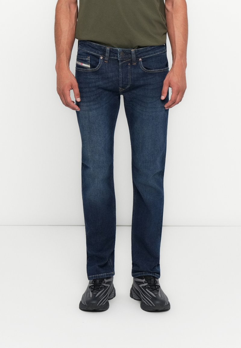 Mann in dunkelblauen Jeans und schwarzen Sportschuhen, der vor einem schlichten hellen Hintergrund steht.