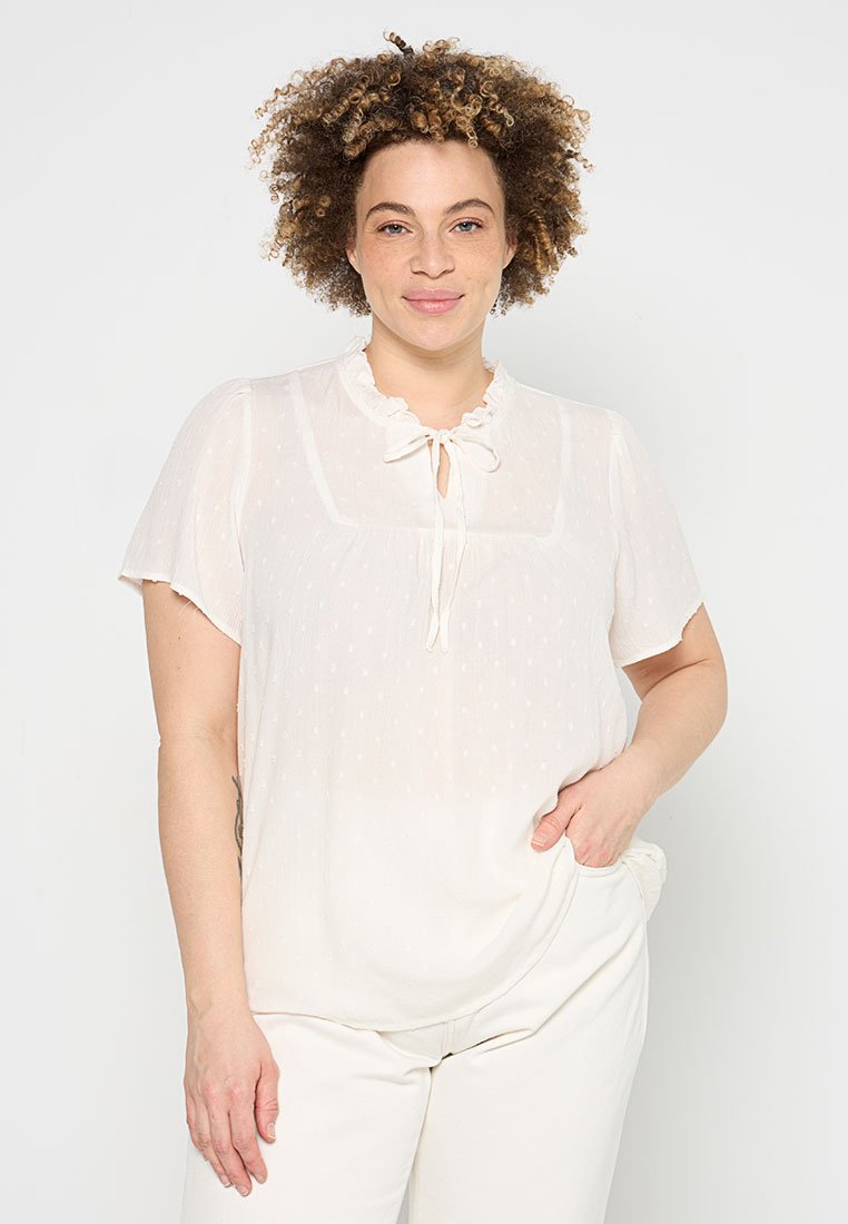 Leichte weiße Bluse mit Rüschenausschnitt, Schleifen-Detail, kurzen Ärmeln und subtiler Strukturierung. Kombiniert mit weißen Hosen.