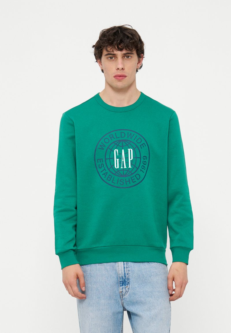 Junger Mann trägt grünes GAP-Sweatshirt mit Globus-Logo und Text „Worldwide Established 1969“, kombiniert mit hellblauen Jeans, vor weißem Hintergrund.