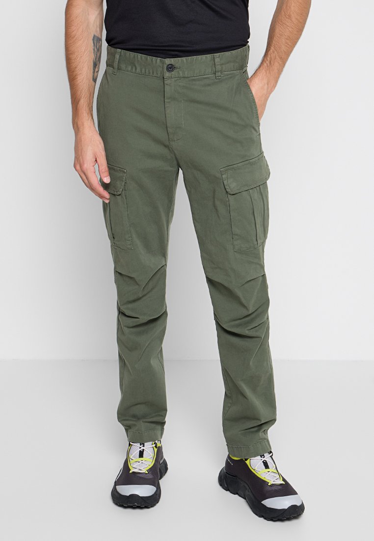 Olivegrüne Cargo-Hosen in lockerer Passform, mit mehreren Seitentaschen, einem Knopfverschluss und einem leichten Stoff. Kombiniert mit schwarzen und gelben Sneakers.