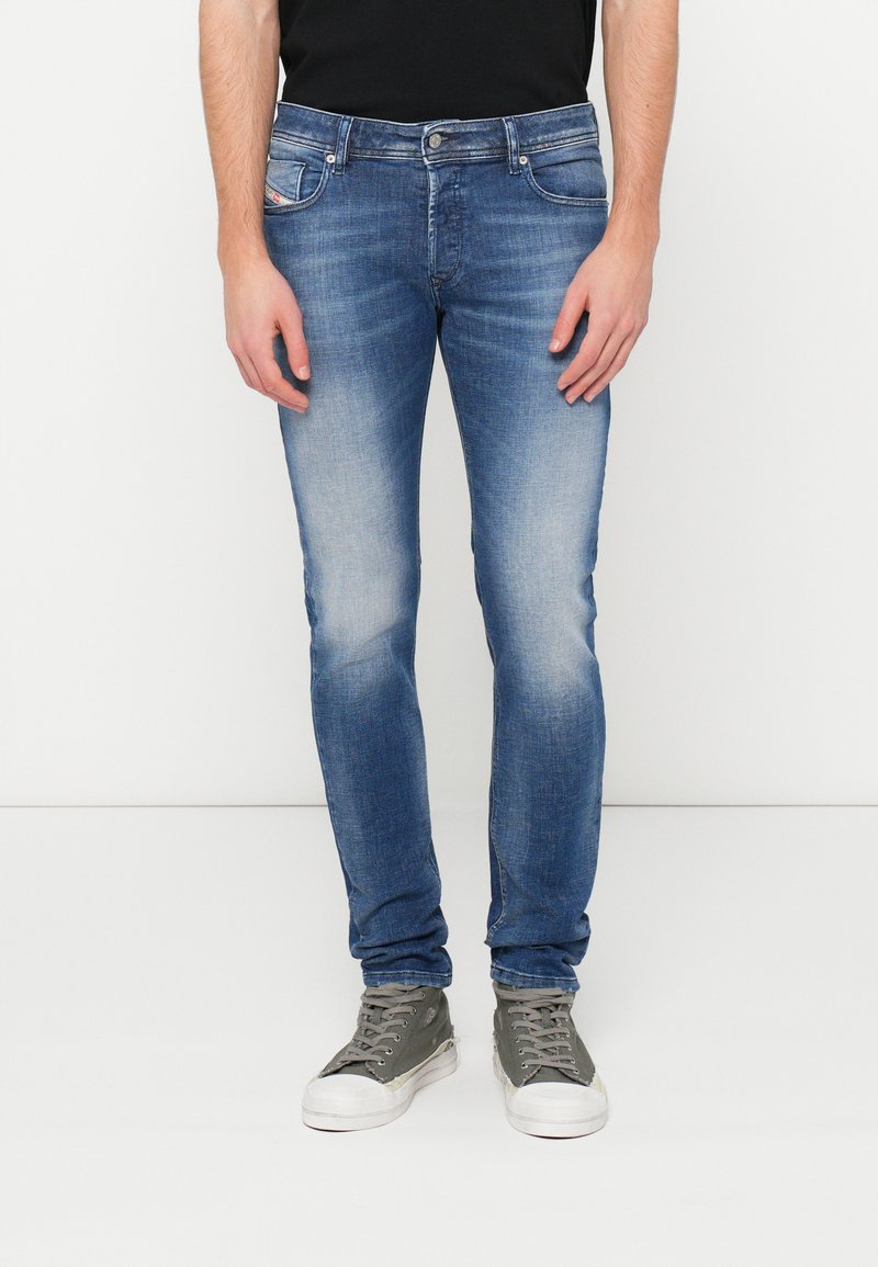 Schmal geschnittene blaue Jeans mit ausgewaschenen Details, fünf Taschen und Knopfverschluss. Kombiniert mit grauen High-Top-Sneakers.