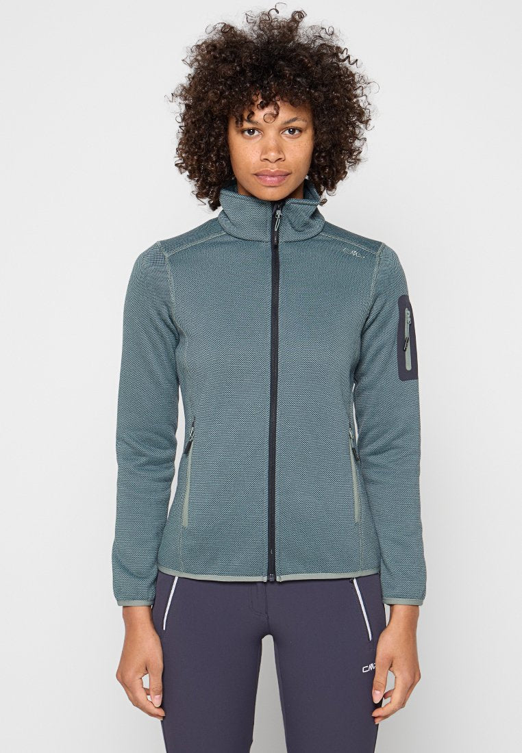 Frau mit lockigem Haar, die eine texturierte blau-graue Jacke mit Reißverschlusstaschen und dunkle, enganliegende Hose vor einem schlichten hellen Hintergrund trägt.