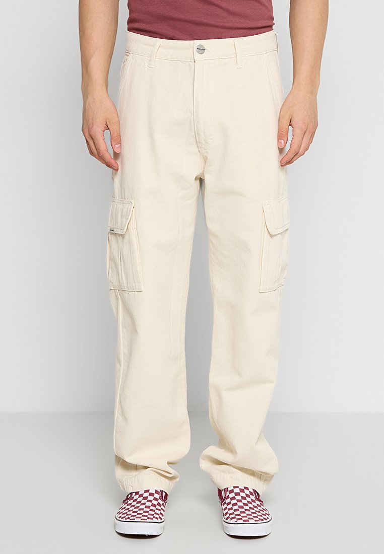 Cremefarbene Cargo-Hosen mit Seitentaschen, lockerer Passform, elastischen Bündchen und einem Metallknopfverschluss. Kombiniert mit rot-weiß karierten Slippern.