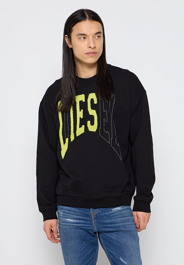 Junger Mann mit langen dunklen Haaren, der einen schwarzen Sweatshirt mit großen gelben und umrandeten Buchstaben sowie eine blaue Jeans trägt und zur Seite schaut.