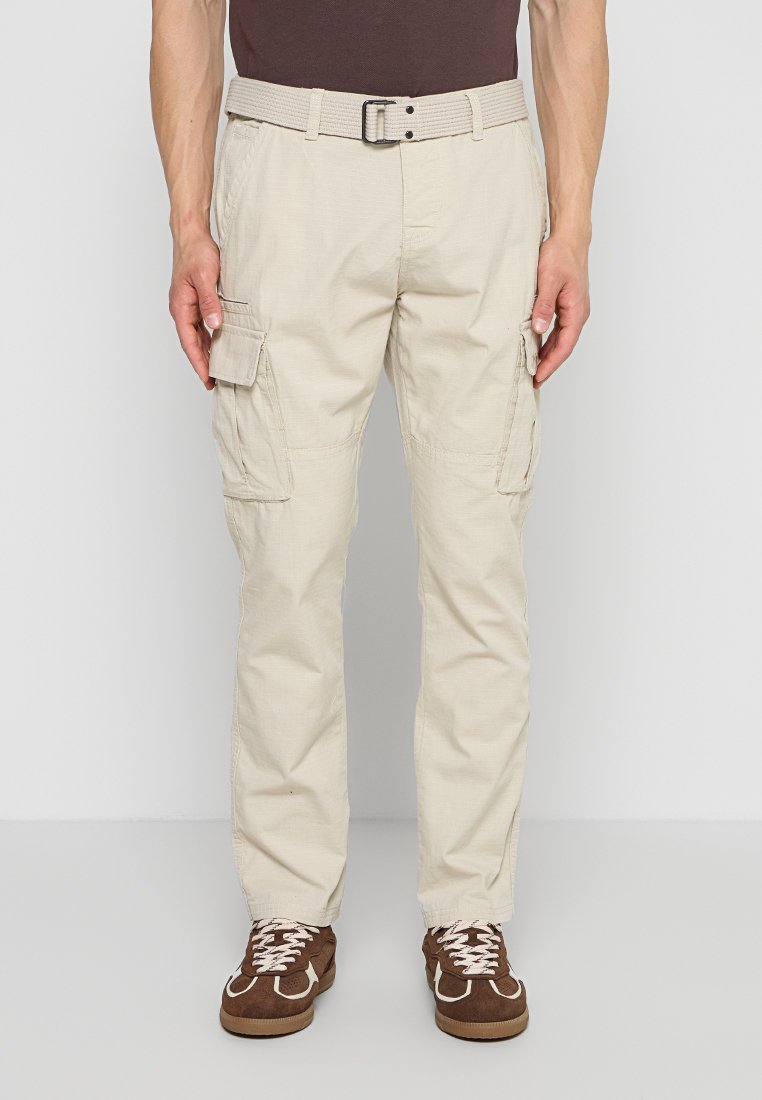 Mann trägt beige Cargohosen mit Seitentaschen, passenden Gürtel, braune Sneaker mit weißen Schnürsenkeln, steht vor einfarbigem Hintergrund.