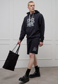 Dunkelgrauer Hoodie mit großem Grafikaufdruck, kombiniert mit passenden Shorts mit kleinem Logo. Schwarze Tragetasche und klobige schwarze Stiefel.