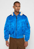 Mann in einer leuchtend blauen Bomberjacke mit weißem Fleecekragen und braunen Hosen, der vor einem schlichten Hintergrund steht.