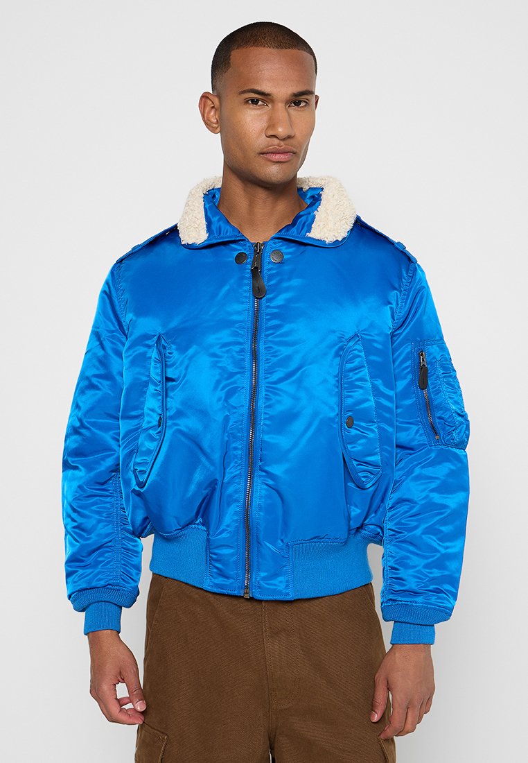 Mann in einer leuchtend blauen Bomberjacke mit weißem Fleecekragen und braunen Hosen, der vor einem schlichten Hintergrund steht.