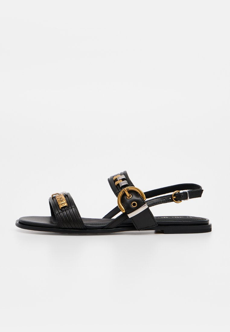 Schwarze flache Sandale mit zwei Riemen, goldenen Schnallendetails und einem Knöchelriemen mit verstellbarem Schnallenverschluss auf weißem Hintergrund.