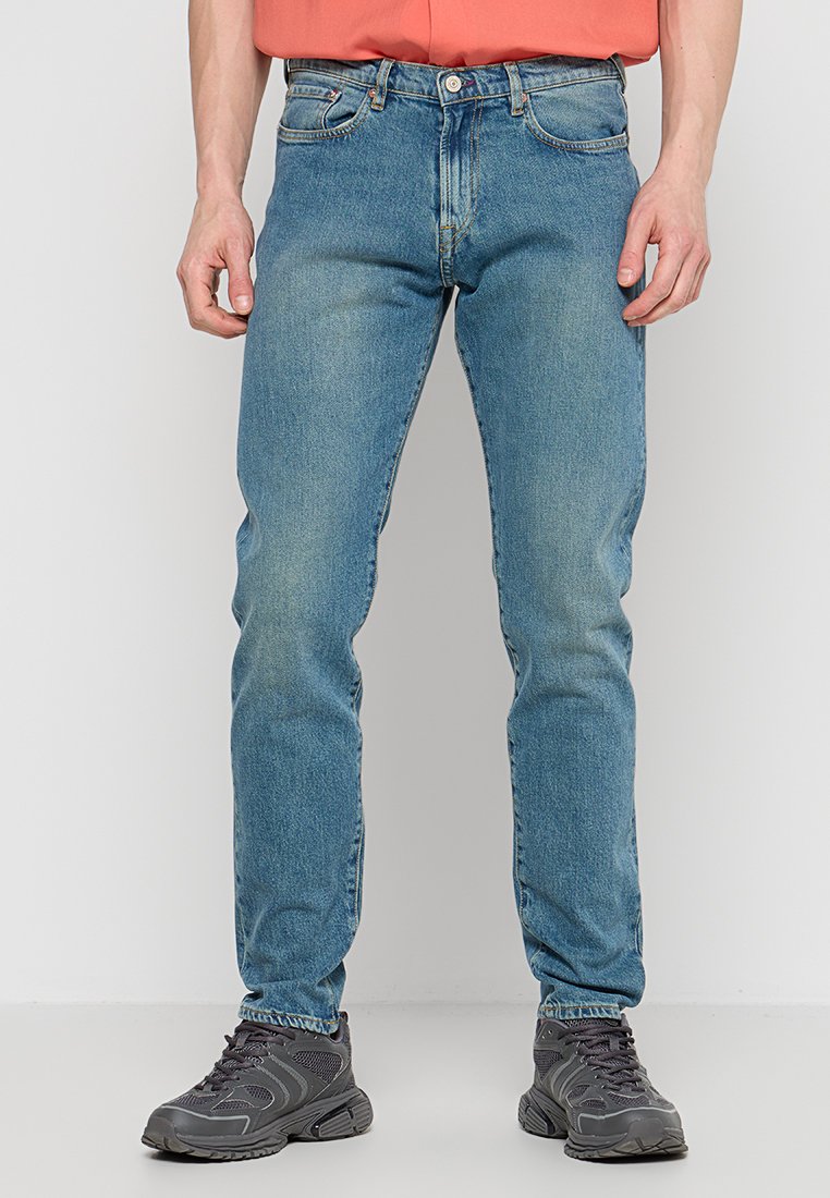 Hellblaue Jeans im fitted Design, klassischer Five-Pocket-Stil und leichter Ausbleichung. Kombiniert mit dunkelgrauen Sportschuhen.