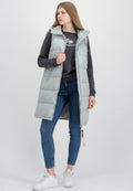 Junge Frau, die eine hellgraue ärmellose Steppweste über einem dunklen Sweatshirt, blaue Skinny Jeans und weiße Sneakers trägt, steht vor einem schlichten Hintergrund.