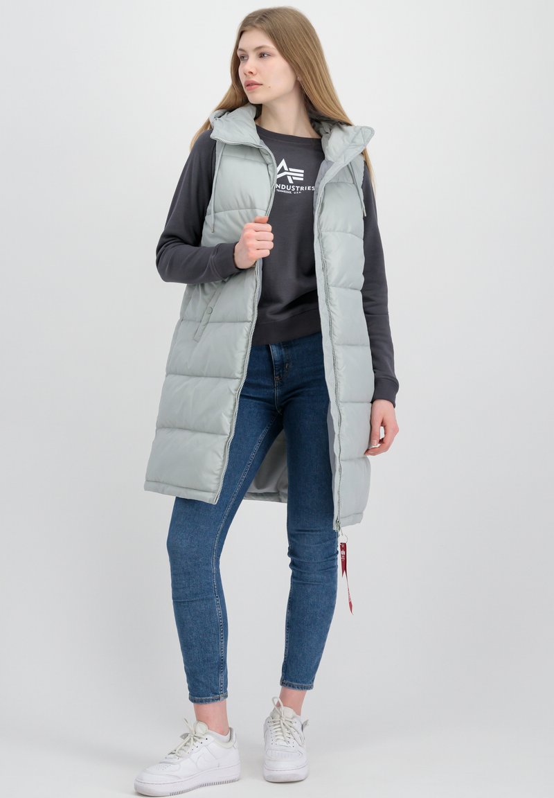 Junge Frau, die eine hellgraue ärmellose Steppweste über einem dunklen Sweatshirt, blaue Skinny Jeans und weiße Sneakers trägt, steht vor einem schlichten Hintergrund.