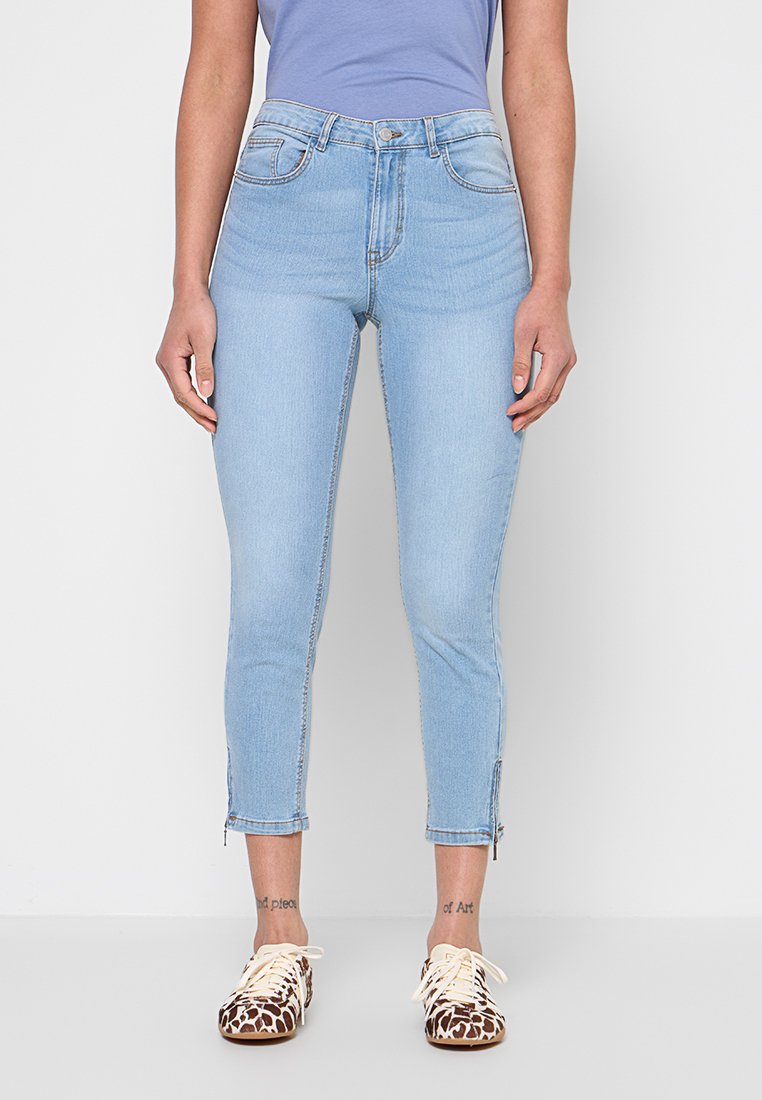 Frau trägt hellblaue, kurze Skinny-Jeans mit Reißverschlüssen am Knöchel, ein in die Hose gestecktes lila Oberteil und Schnürschuhe mit Tiermuster.