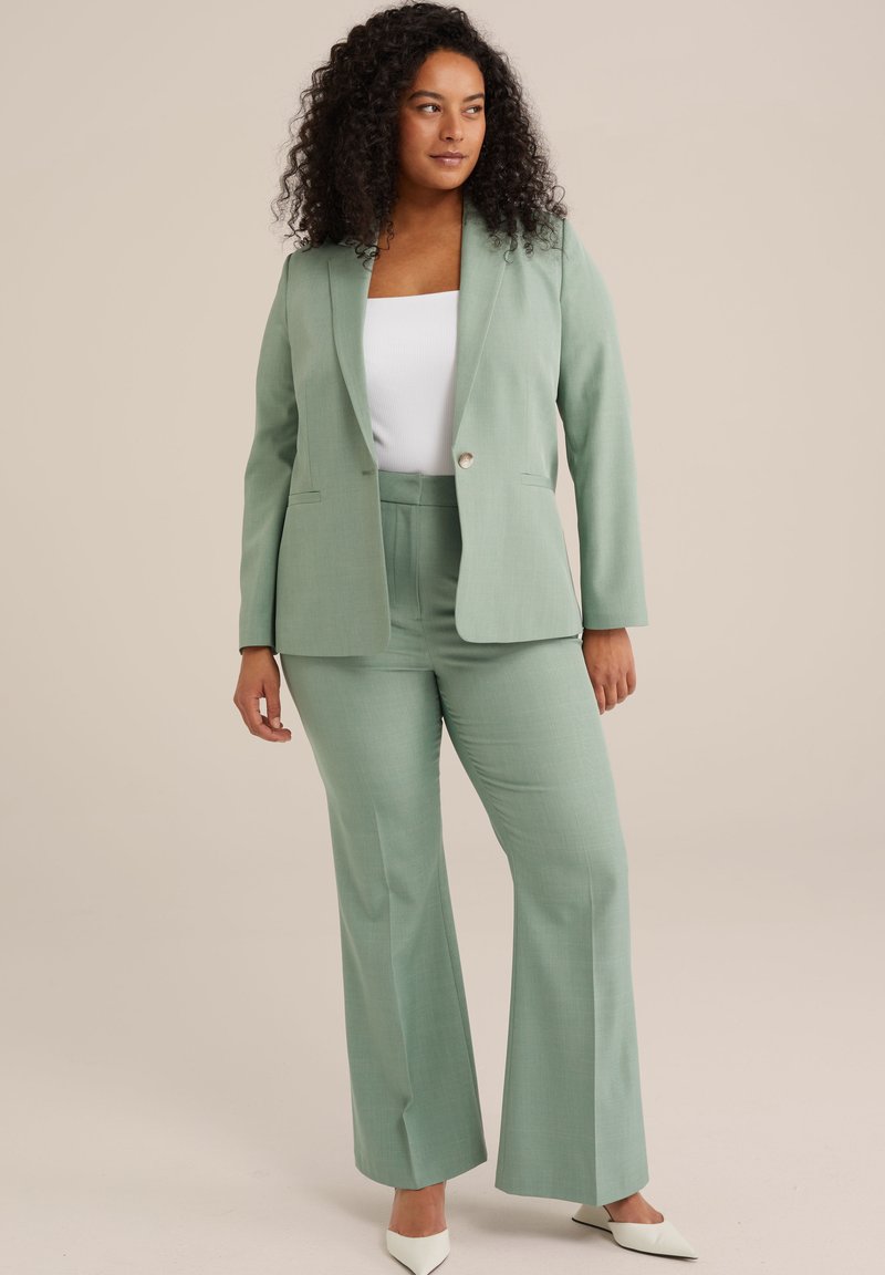 Hellegrüner Anzug mit maßgeschneidertem Blazer und ausgestellten Hosen. Glatte Textur, ein einzelner Knopf vorne und Seitentaschen. Weiße High Heels.