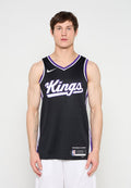 Junger Mann, der ein schwarzes Sacramento Kings Basketballtrikot und weiße Shorts trägt, steht vor einem weißen Hintergrund und blickt nach vorne.