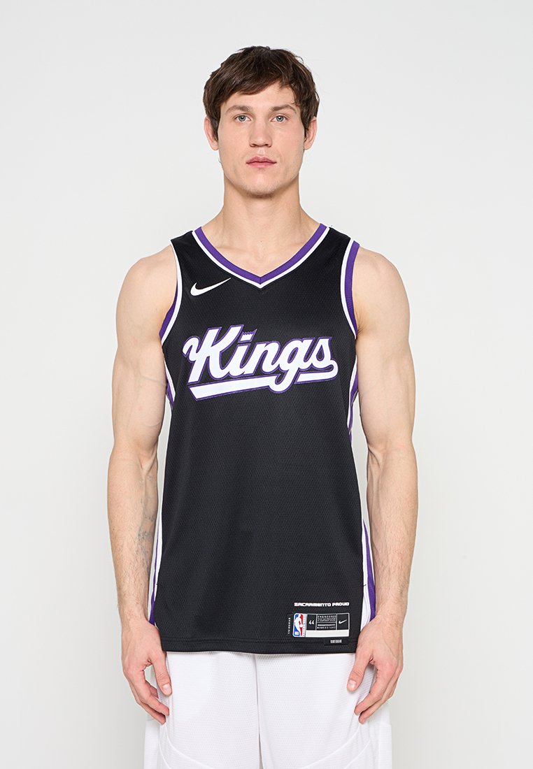 Junger Mann, der ein schwarzes Sacramento Kings Basketballtrikot und weiße Shorts trägt, steht vor einem weißen Hintergrund und blickt nach vorne.
