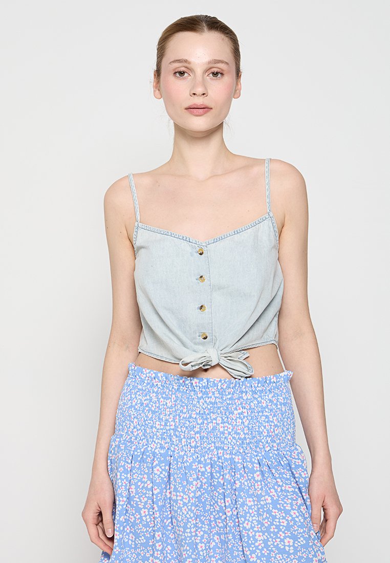 Junge Frau mit gebundenem, hellem Jeanshemd und hohem blumenförmigen Rock in Blau vor einem schlichten, hellen Hintergrund.