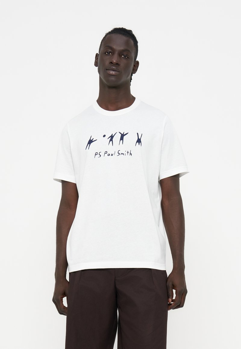 Mann trägt ein weißes T-Shirt mit schwarzen abstrakten Figuren und dem Schriftzug „PS Paul Smith“, kombiniert mit dunkelbrauner Hose, steht vor weißem Hintergrund.