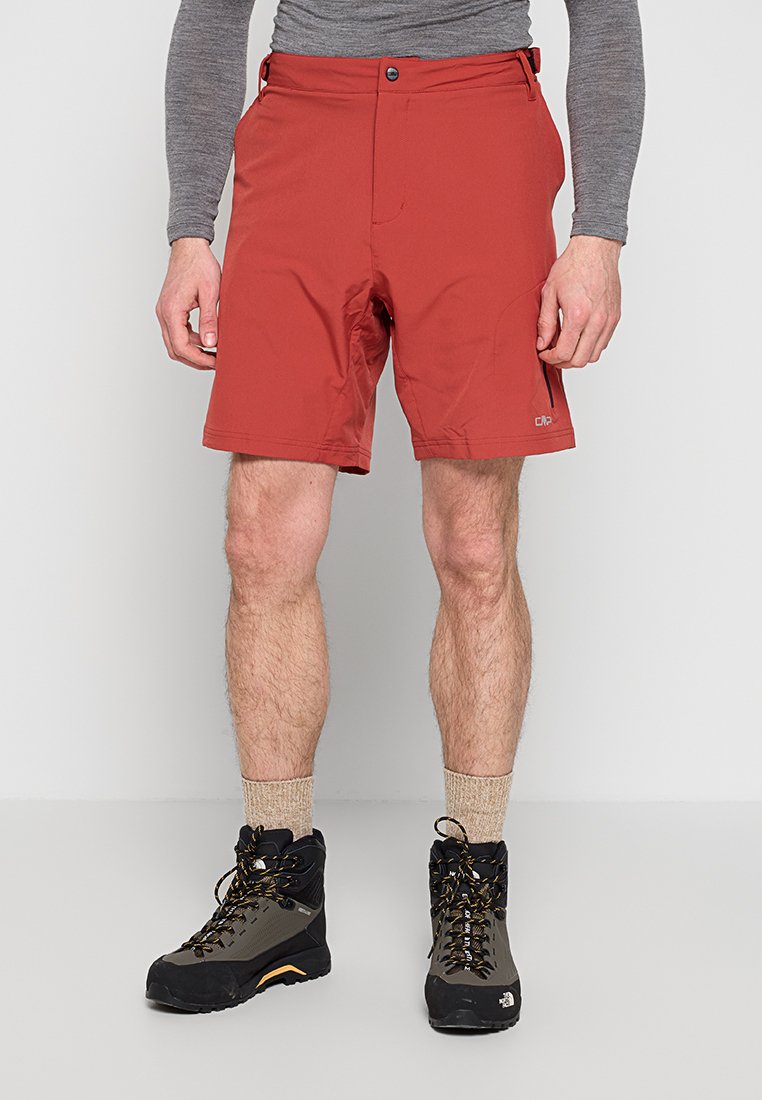 Mann trägt rote Wandershorts, beige Socken und schwarze hochgeschnittene Wanderschuhe, steht vor einem einfarbigen Hintergrund.