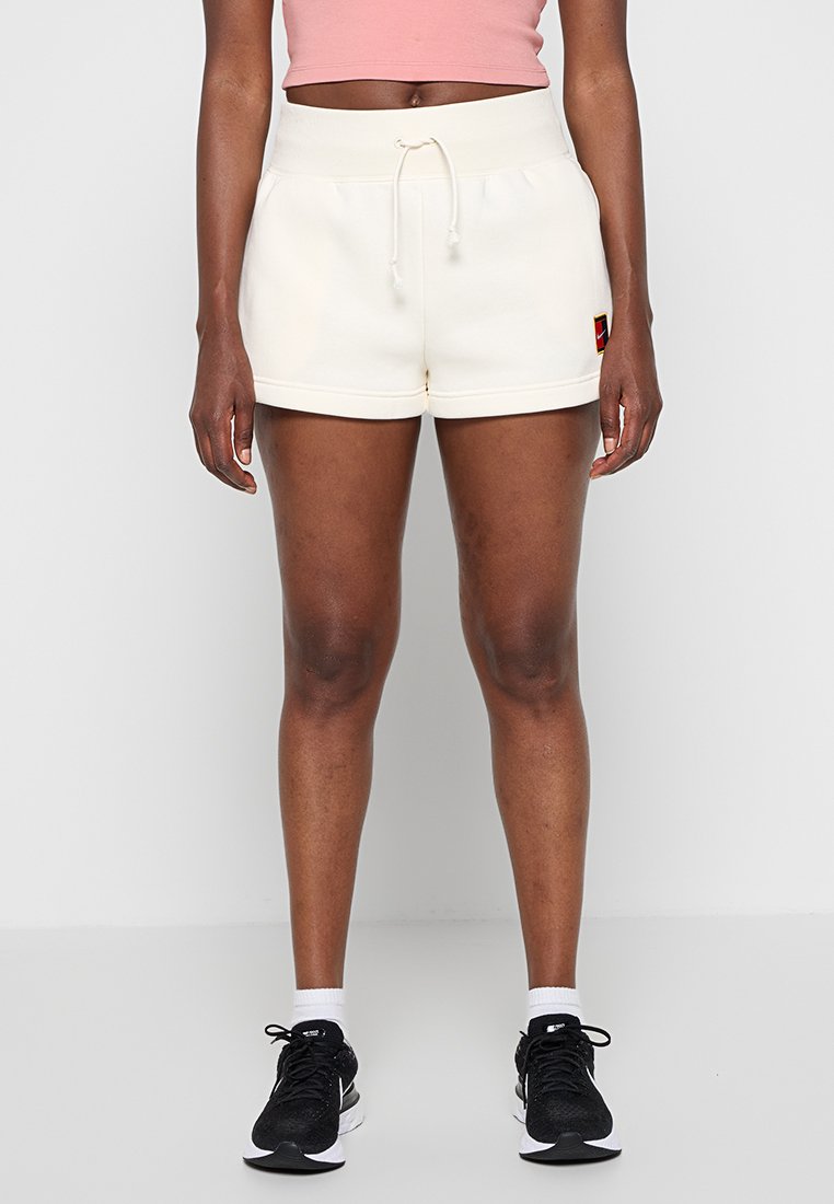 Person trägt cremefarbene Shorts mit Kordelzug, ein pinkes Croptop, weiße Socken und schwarze Sneakers und steht vor einem schlichten grauen Hintergrund.