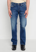 Mann trägt ausgewaschene blaue Jeans mit geradem Bein und schwarze Lederschuhe, steht vor einem einfarbigen hellen Hintergrund.