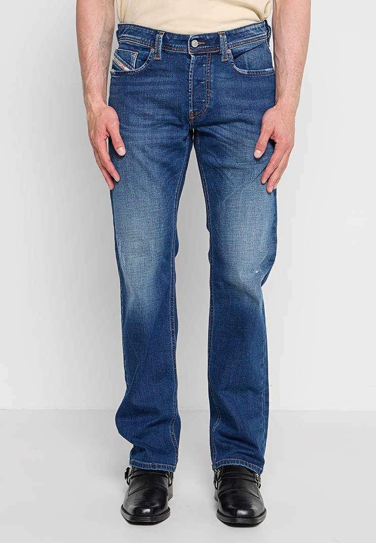 Mann trägt ausgewaschene blaue Jeans mit geradem Bein und schwarze Lederschuhe, steht vor einem einfarbigen hellen Hintergrund.