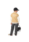 Hellbraune und weiße Baseballjacke mit der Aufschrift "Athletics Club", schwarze Kappe, graue Jeanshose und schwarze Tasche mit weißem Logo. Die Sneakers haben gedämpfte Farben.