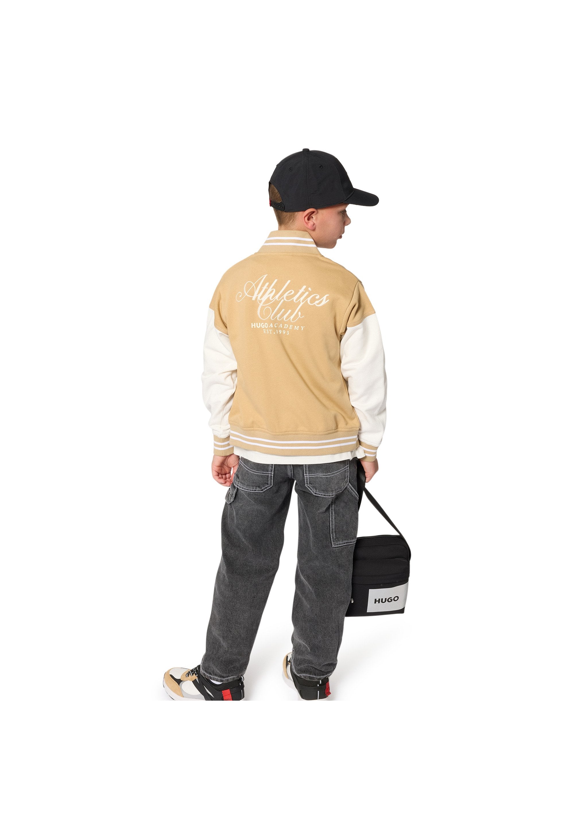 Hellbraune und weiße Baseballjacke mit der Aufschrift "Athletics Club", schwarze Kappe, graue Jeanshose und schwarze Tasche mit weißem Logo. Die Sneakers haben gedämpfte Farben.
