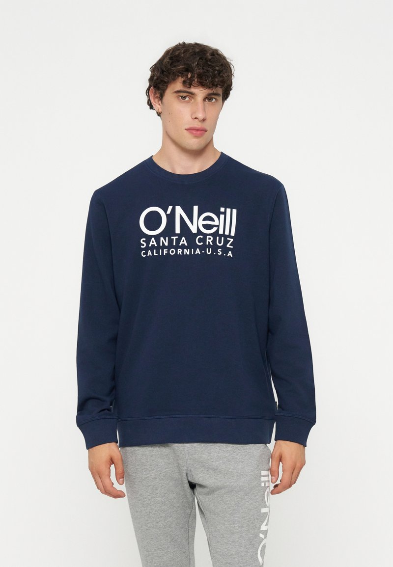 Junger Mann mit lockigem Haar, der einen marineblauen O'Neill-Sweatshirt und graue Sweatpants mit Logo trägt, steht vor einem weißen Hintergrund.