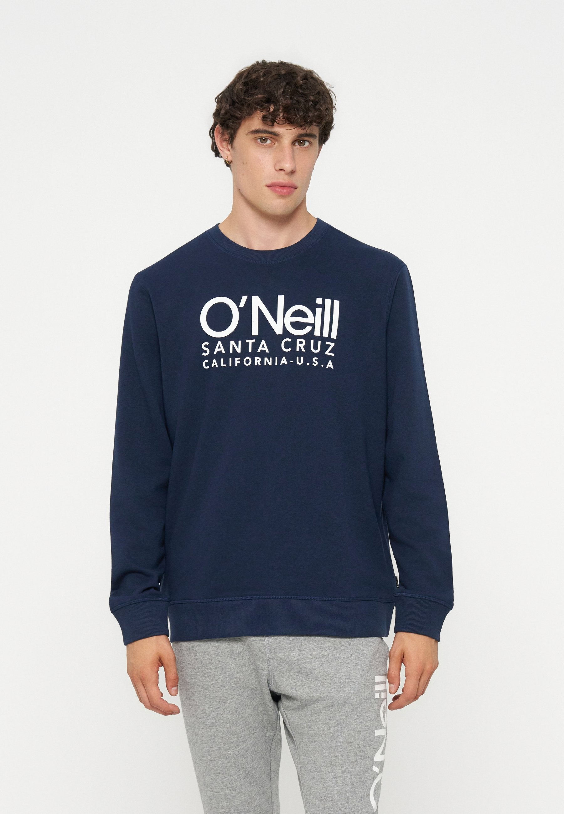 Junger Mann mit lockigem Haar, der einen marineblauen O'Neill-Sweatshirt und graue Sweatpants mit Logo trägt, steht vor einem weißen Hintergrund.