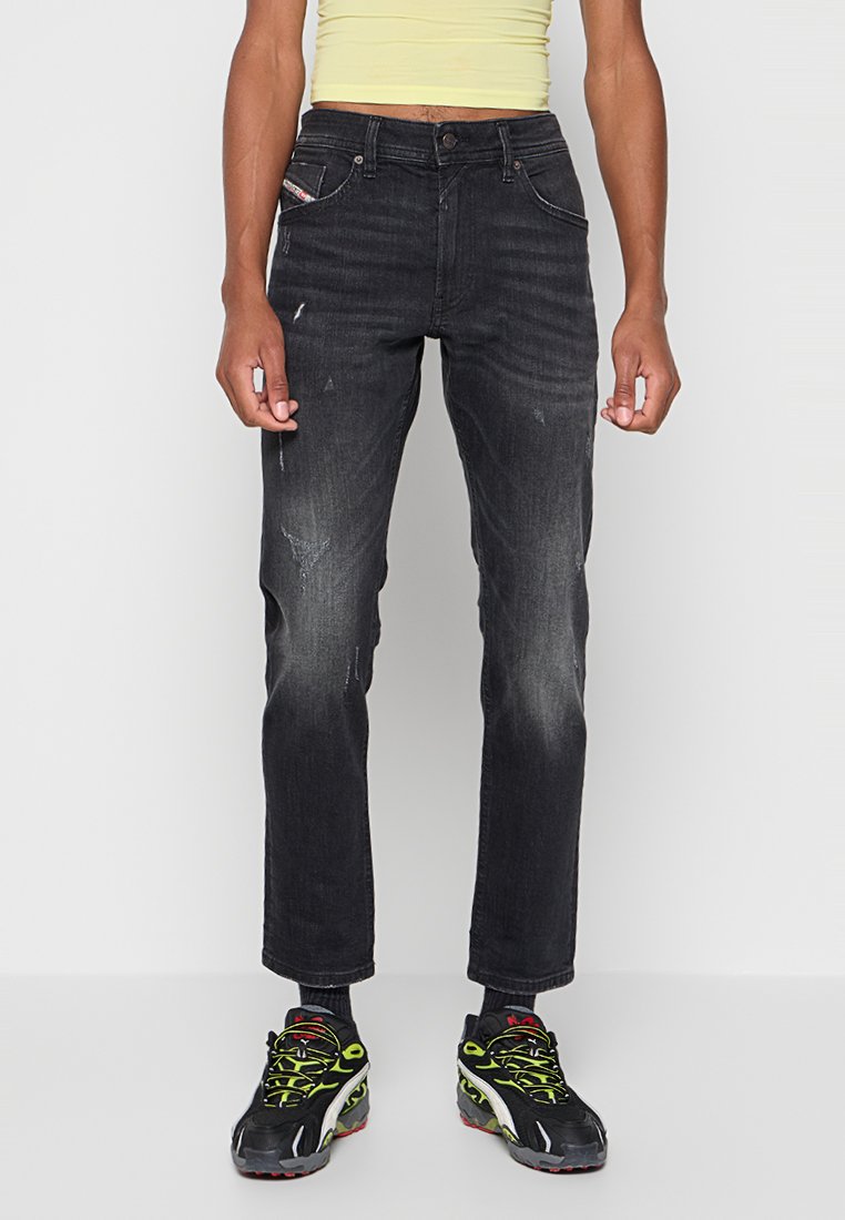 Person, die verblasste schwarze Jeans mit Used-Effekten, schwarze Sneakers mit neongrünen Schnürsenkeln und ein hellgelbes Crop-Top trägt, steht vor einem weißen Hintergrund.