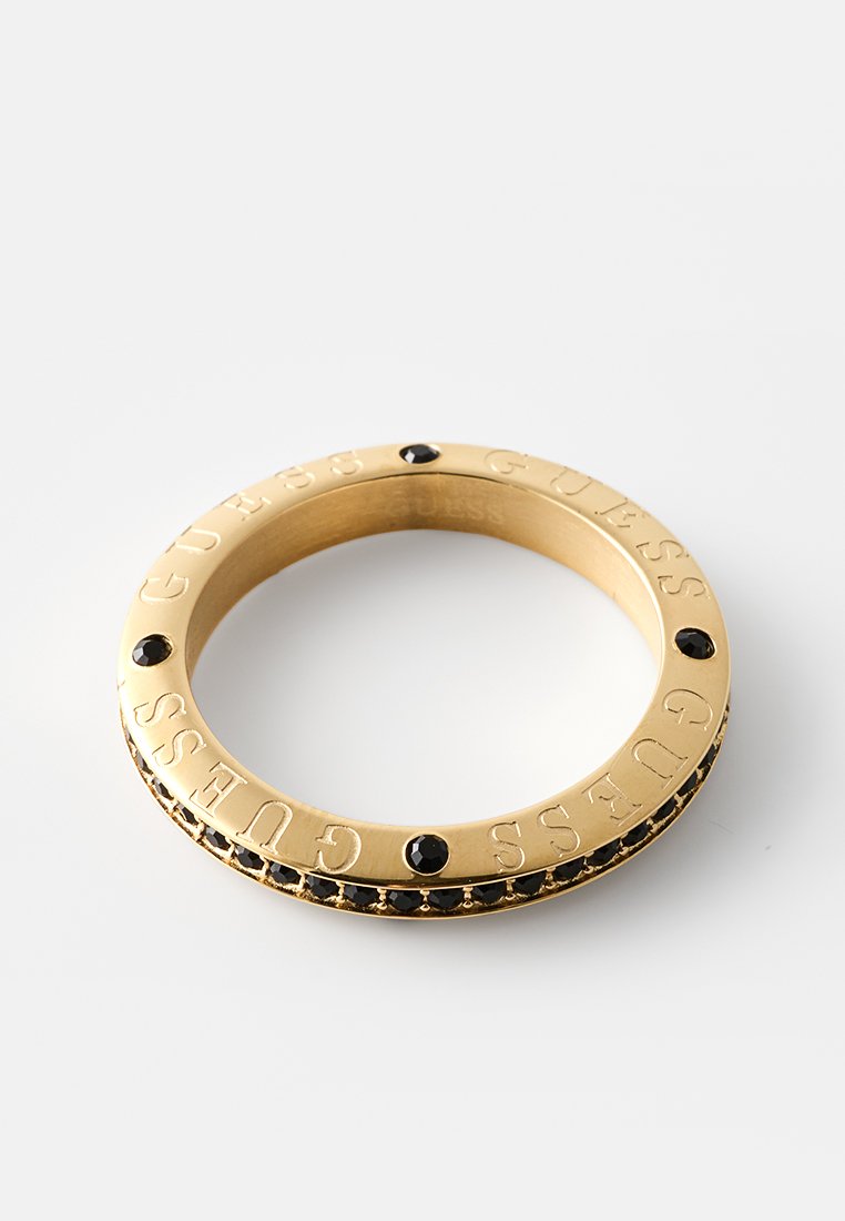 Goldener Armreif mit eingraviertem "GUESS"-Schriftzug, versehen mit schwarzen Akzenten und Steinen rund um das Band. Glatte Textur und rundes Design.