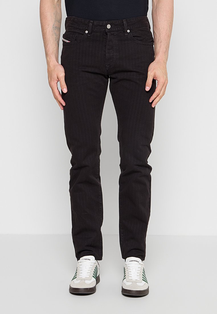 Mann trägt schlanke schwarze Jeans und weiße Sneaker mit grünen Streifen, steht mit entspannten Händen an den Seiten vor einfarbigem Hintergrund.