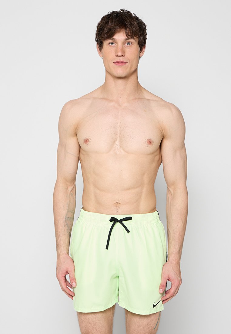 Oberkörperfreier Mann mit kurzen dunklen Haaren, der hellgrüne Nike-Badeshorts mit schwarzem Kordelzug trägt und vor einem einfarbigen grauen Hintergrund steht.