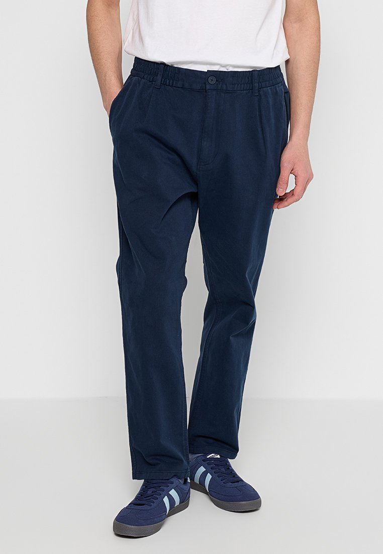 Marineblaue Baumwollhose mit elastischem Bund, seitlichen Taschen und lockerer Passform. Kombiniert mit marineblauen Turnschuhen mit hellblauen Streifen.