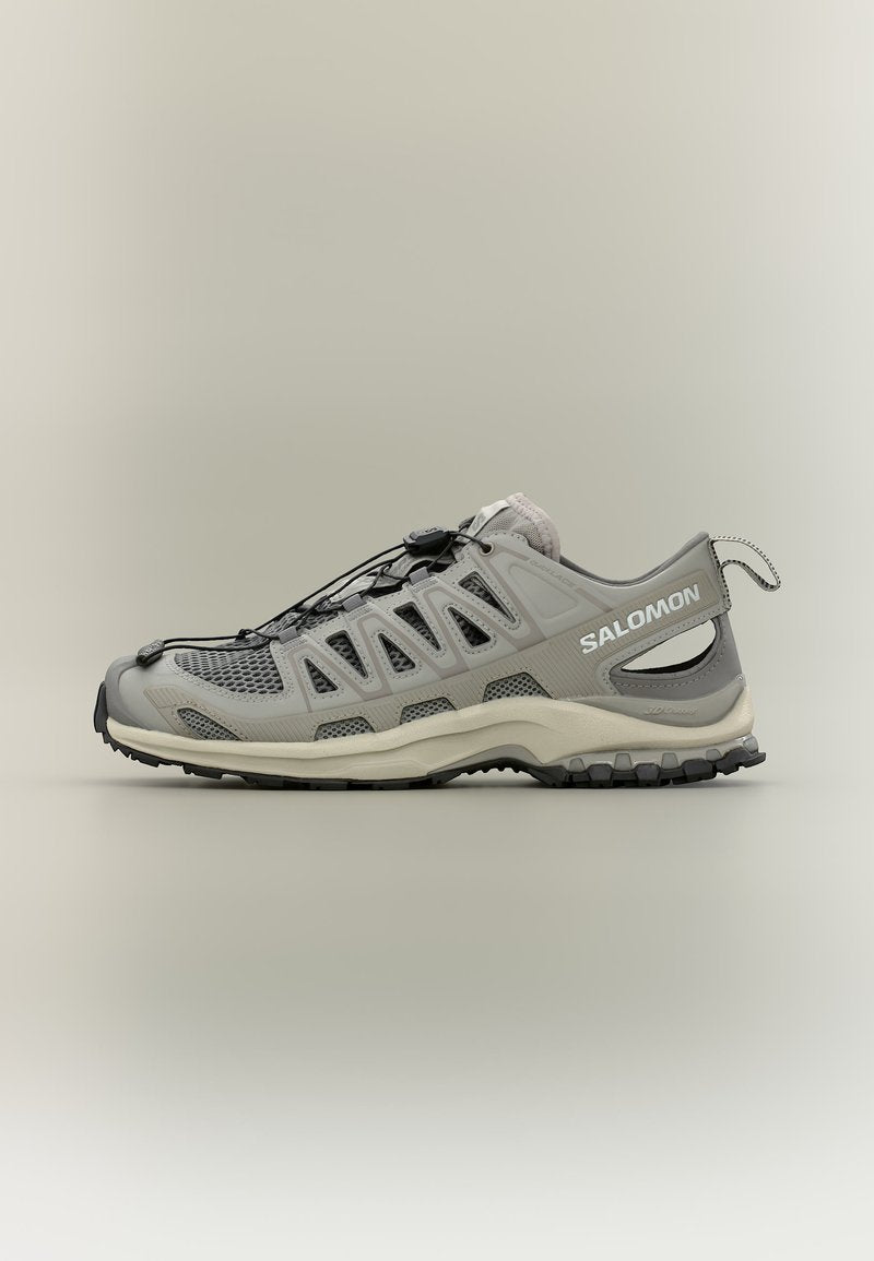 Hellgrauer Salomon Trailrunning-Schuh mit Mesh-Einsätzen, Schnellbändersystem und robuster schwarzer Sohle, seitlich auf neutralem Hintergrund gezeigt.